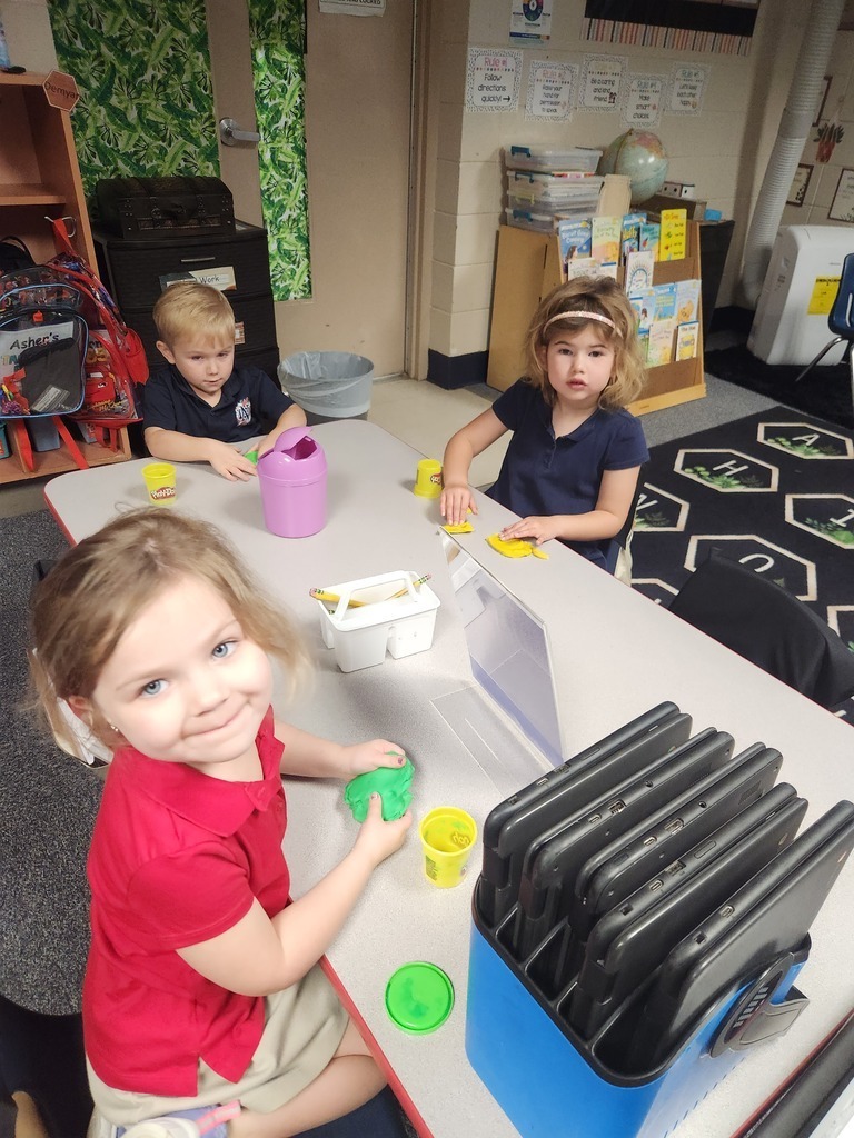 Three children, including a smiling girl, sit at a table with various objects, such as a purple bucket, a yellow container, and a blue box.