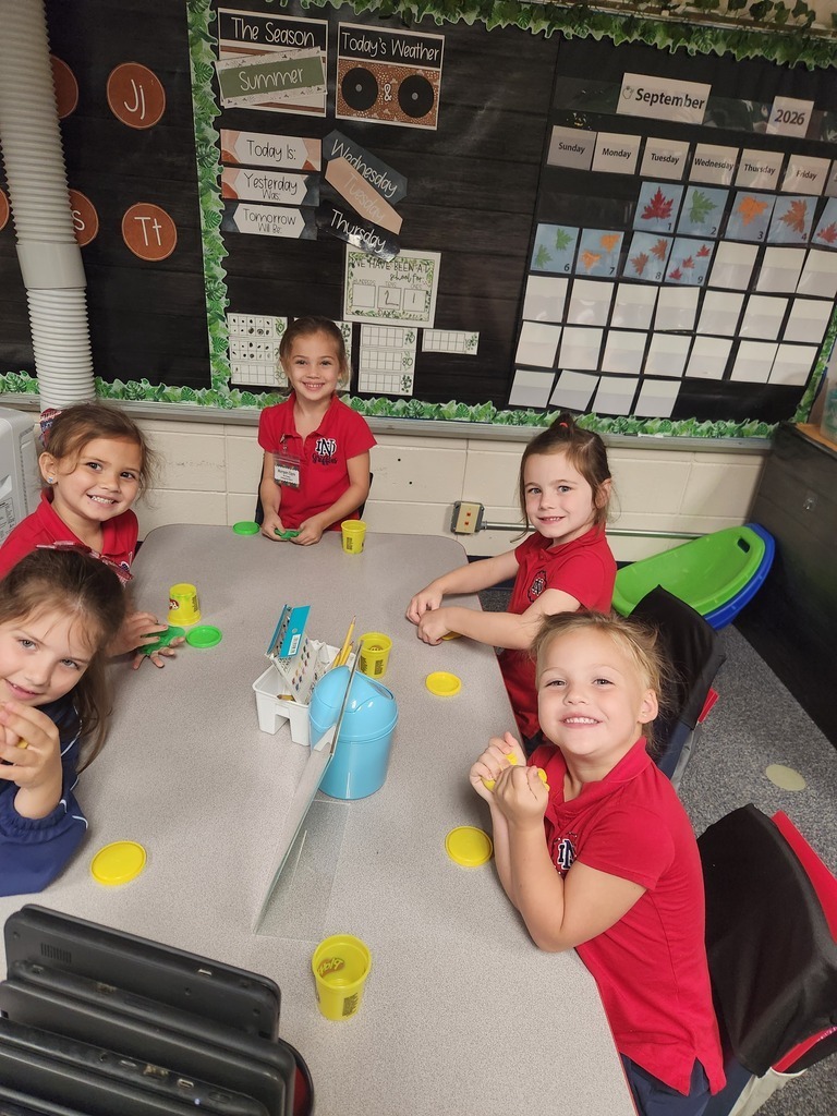 Five young children wearing red shirts sit at a table with yellow cups and a blue bucket.