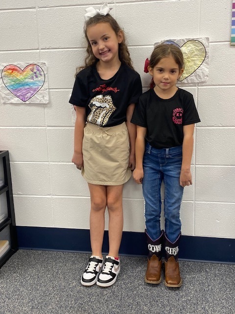 Two children, one with a skirt and the other in jeans, stand next to a wall with heart-shaped artwork.