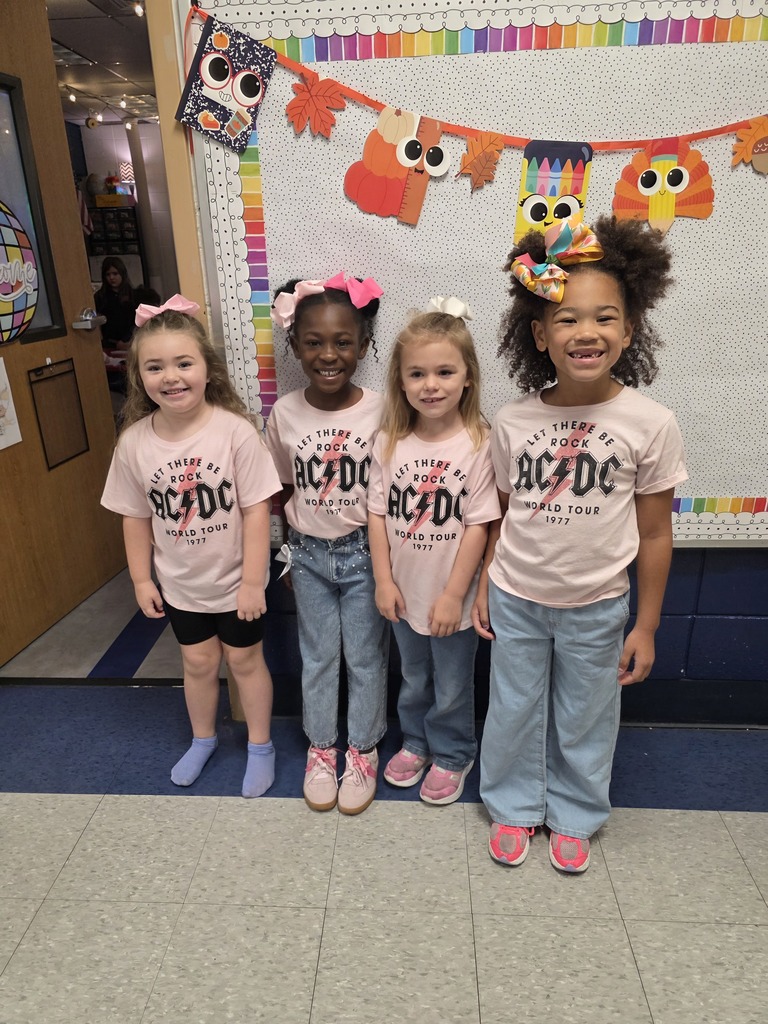 Four girls wearing pink AC/DC shirts and various colored shoes stand together in front of a colorful banner.