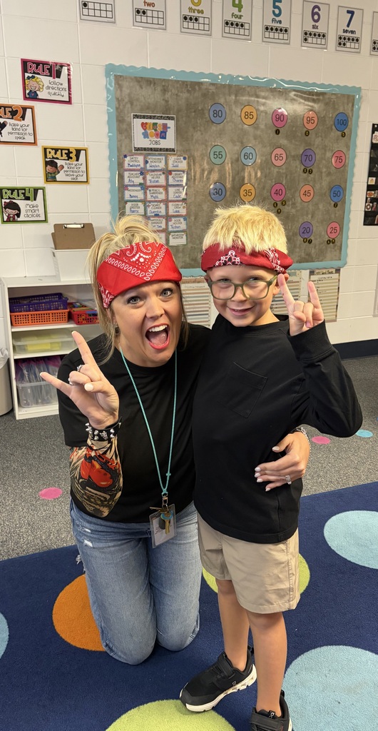 A woman and a boy crouch on a multicolored floor, smiling and making peace signs. The woman wears a red bandana, and the boy wears glasses. Behind them is a wall with numbers and cards.