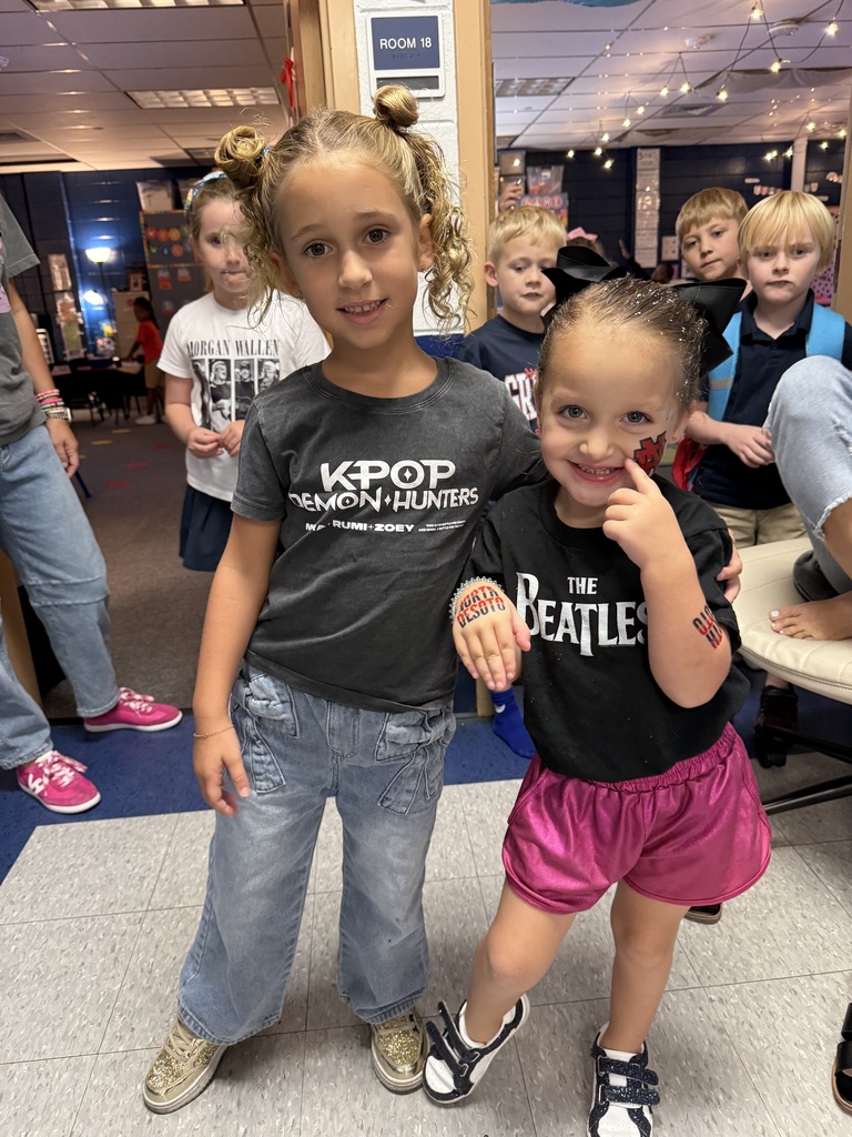 Two young girls with smiling faces stand close together in a room with other children.