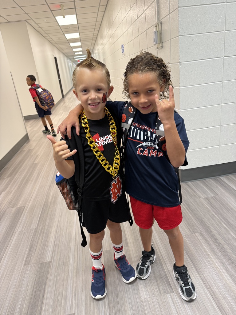 Two young boys pose in a hallway. One with a backpack and mohawk, the other in red shorts.