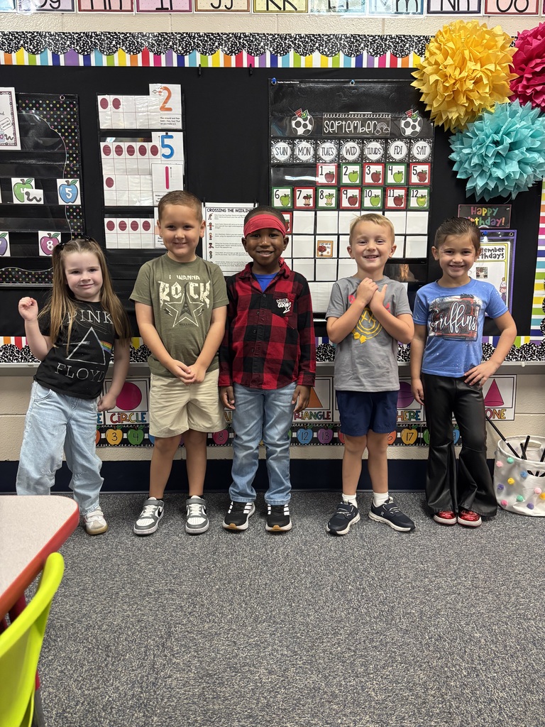 Five children in a classroom with a bulletin board and pom-poms. They stand in a line.