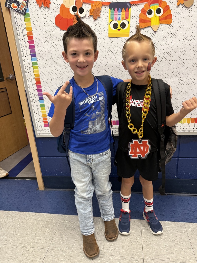 Two boys in school uniforms, one wearing a blue shirt and the other a black shirt, standing in a classroom with decorations in the background.