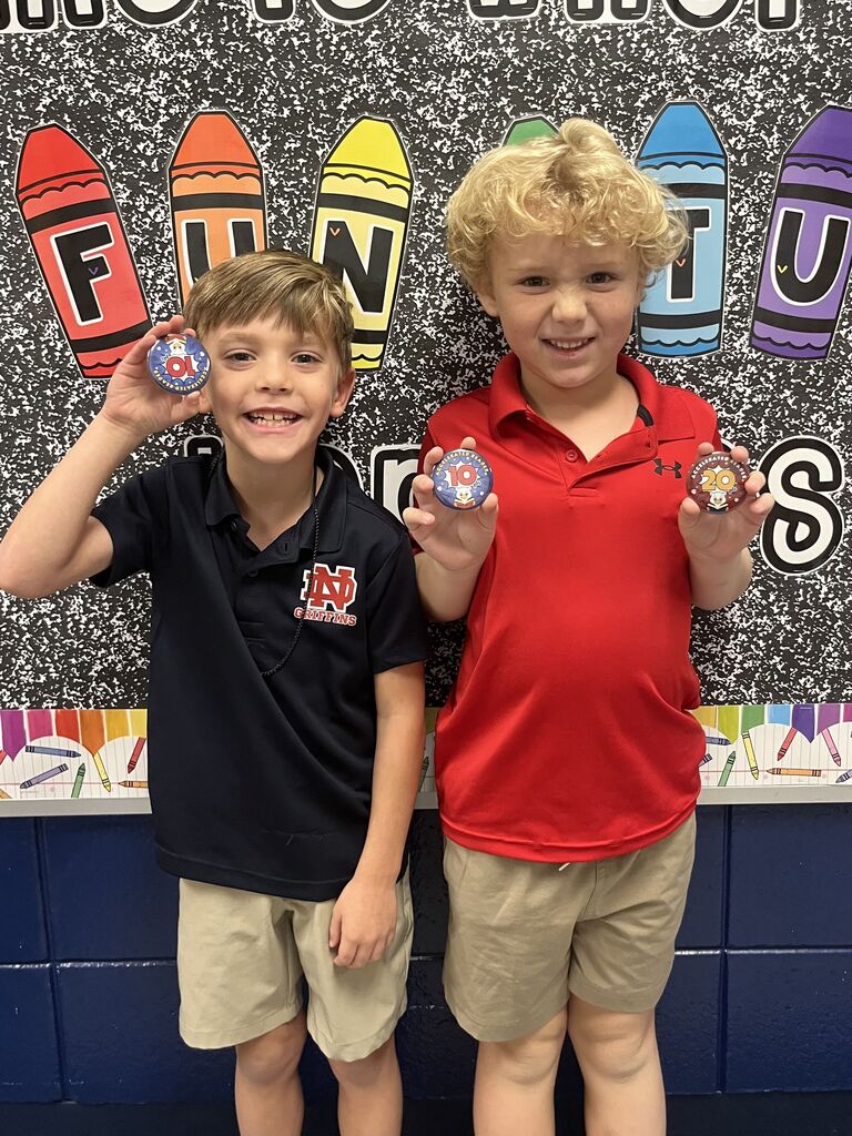 Two boys stand together, smiling and holding small objects. One wears a blue polo shirt; the other, red. Behind them is a colorful, educational wall with letters.