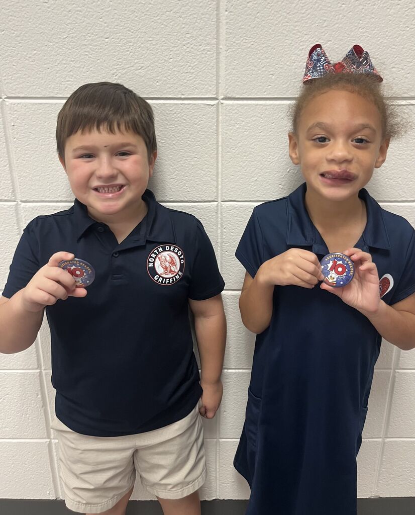 Two children in navy blue uniforms, standing next to each other. One holds a pin, and the other holds a medal.