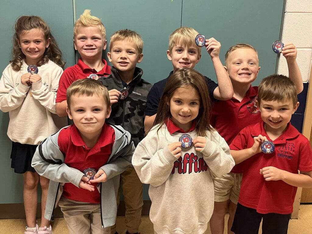 A group of children in a room, each wearing red shirts, holding medals. A girl in a white shirt and shorts stands at the left.