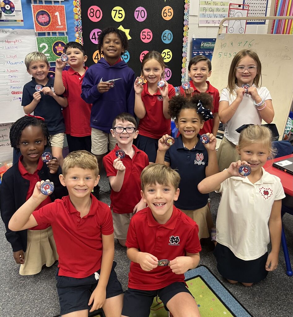 Elementary students wearing red and blue shirts and white tops hold small plastic objects in their hands.
