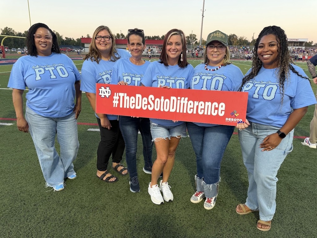 Six women in blue shirts and jeans hold a red sign with "#TheDeSotoDifference" on a sports field.