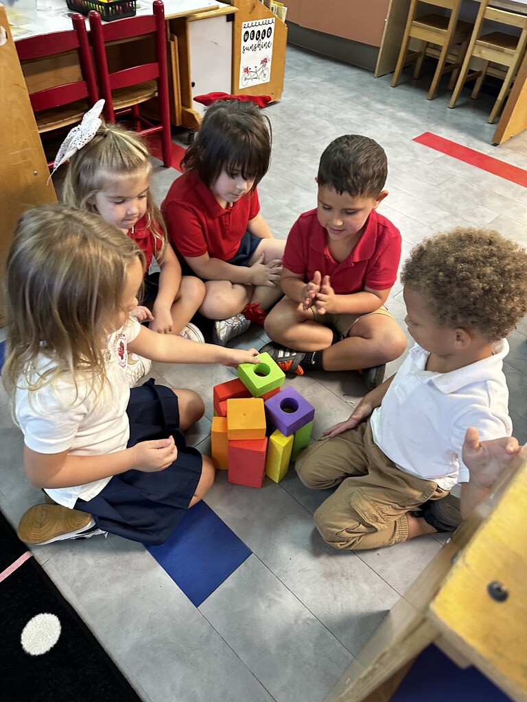 Several young children sit on a floor, playing with colorful cubes. The room has a simple setup with chairs and desks.
