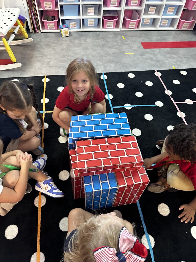 Group of children sitting on floor around a large multi-colored cube, with storage bins in background.
