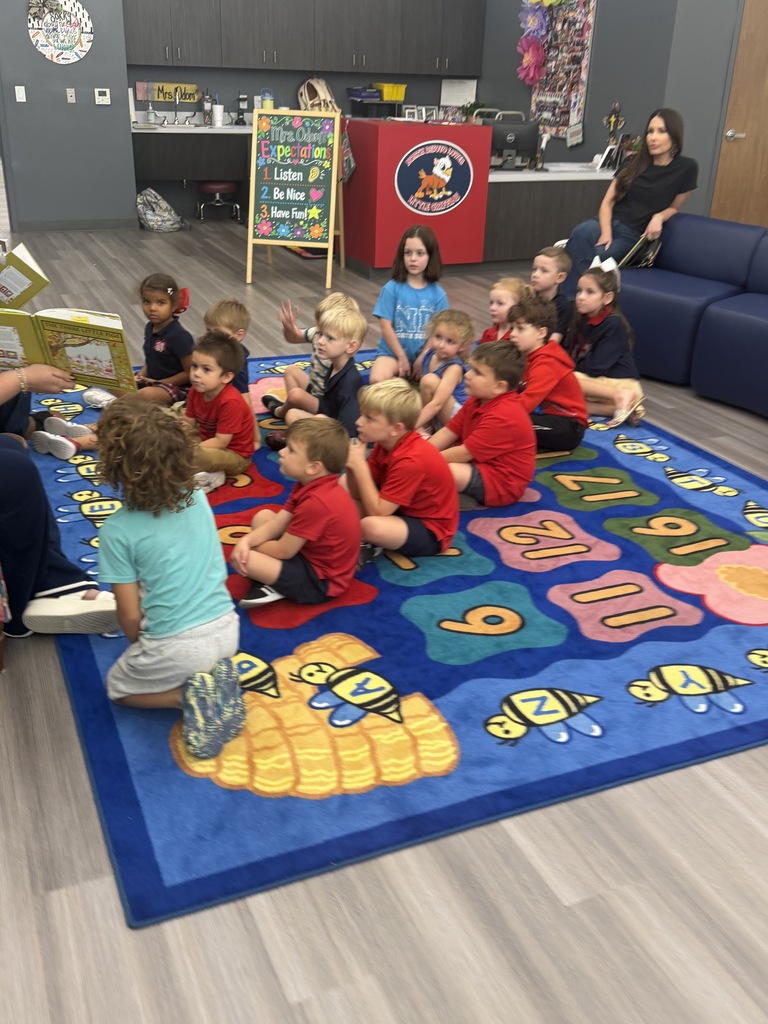 A classroom with children sitting in a circle on a colorful mat, listening to a person read aloud.
