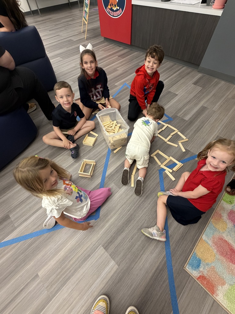 Children sit around a table and floor, playing with building blocks. A couch and a colorful rug are in the background.