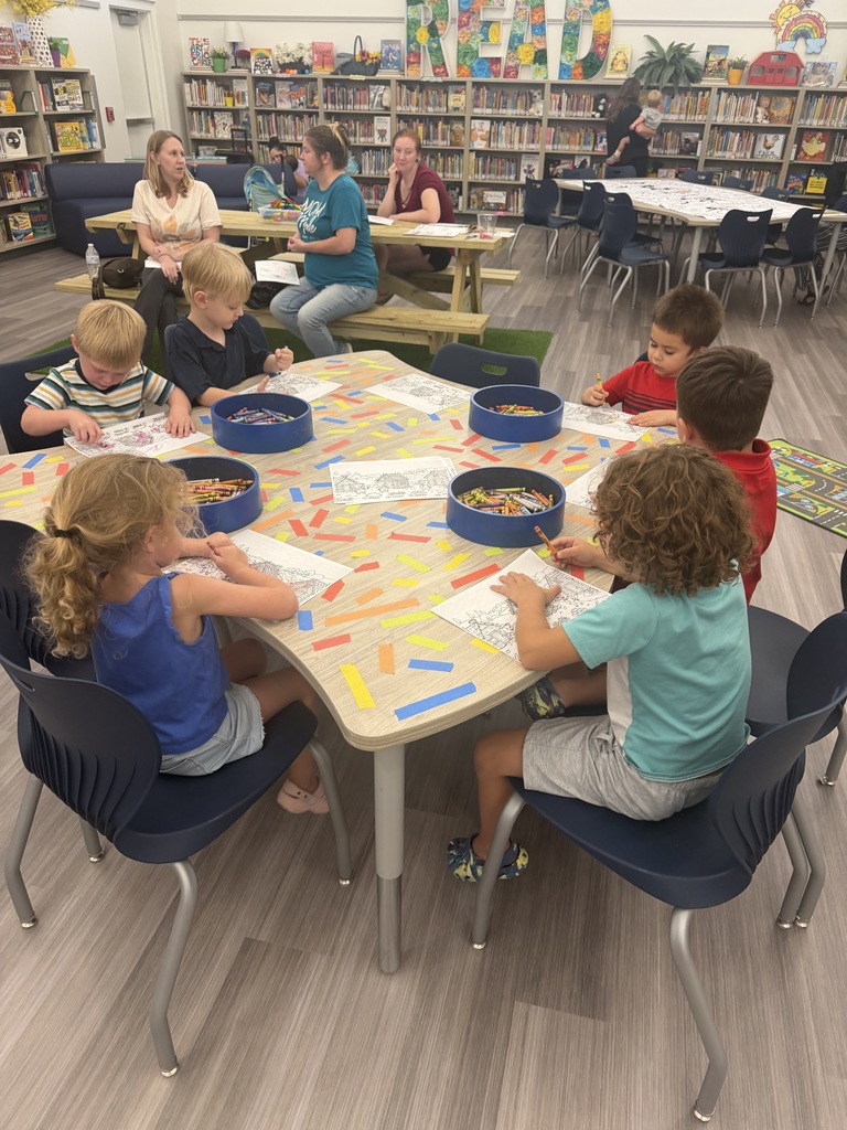 Kids sit around a table in a library. They are doing arts and crafts with colored paper.