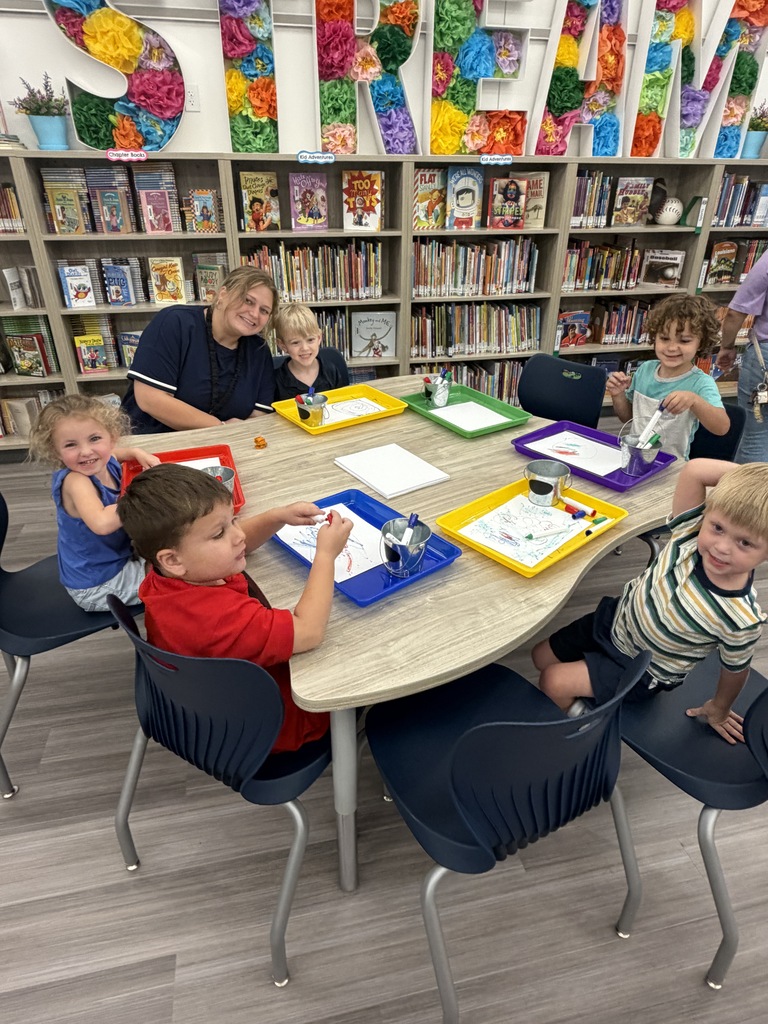 Children seated at a table, working on crafts in a library with shelves filled with books.