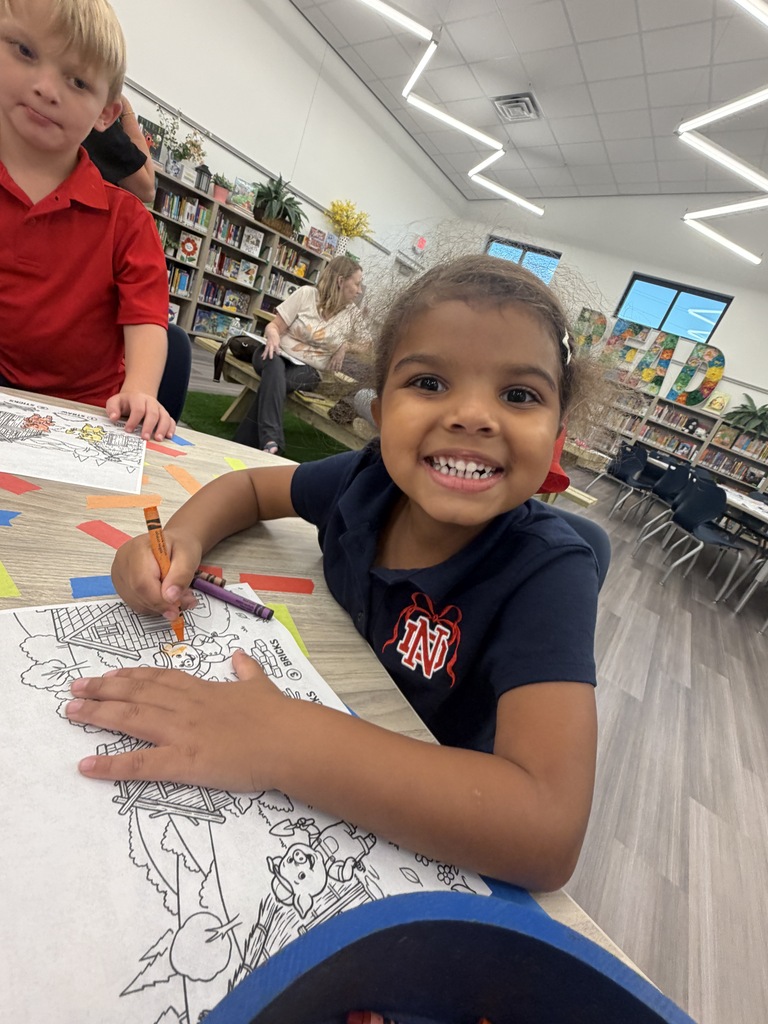 A young girl smiles while coloring a picture in a classroom. A boy sits behind her, also coloring.