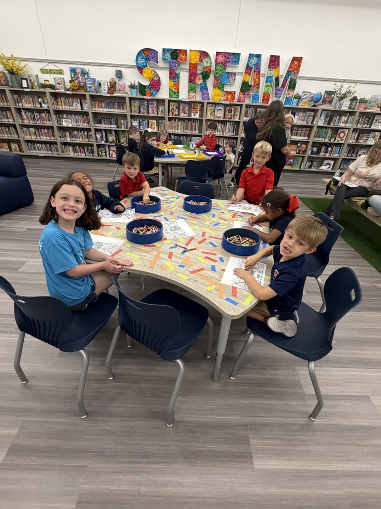 Kids seated around a table in a library, drawing on paper with crayons. Library shelves in the background.