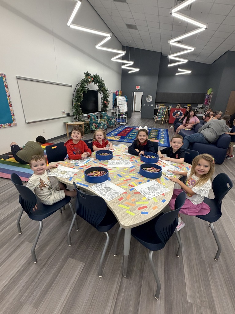 Kids sit around a table with papers and activities in a room with blue chairs and a wooden floor.