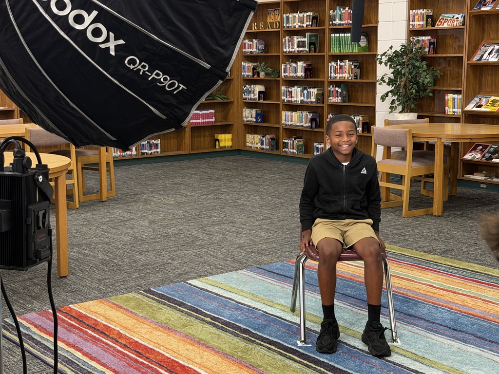 A child sits in a chair with a light behind them in a library. Colorful rug and wooden bookshelves.