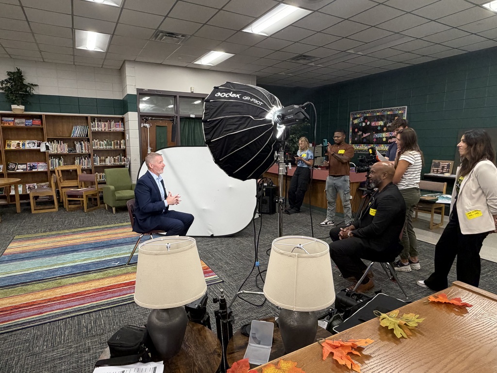 Indoor room with people setting up equipment for a video shoot. A man sits on a chair in the middle.