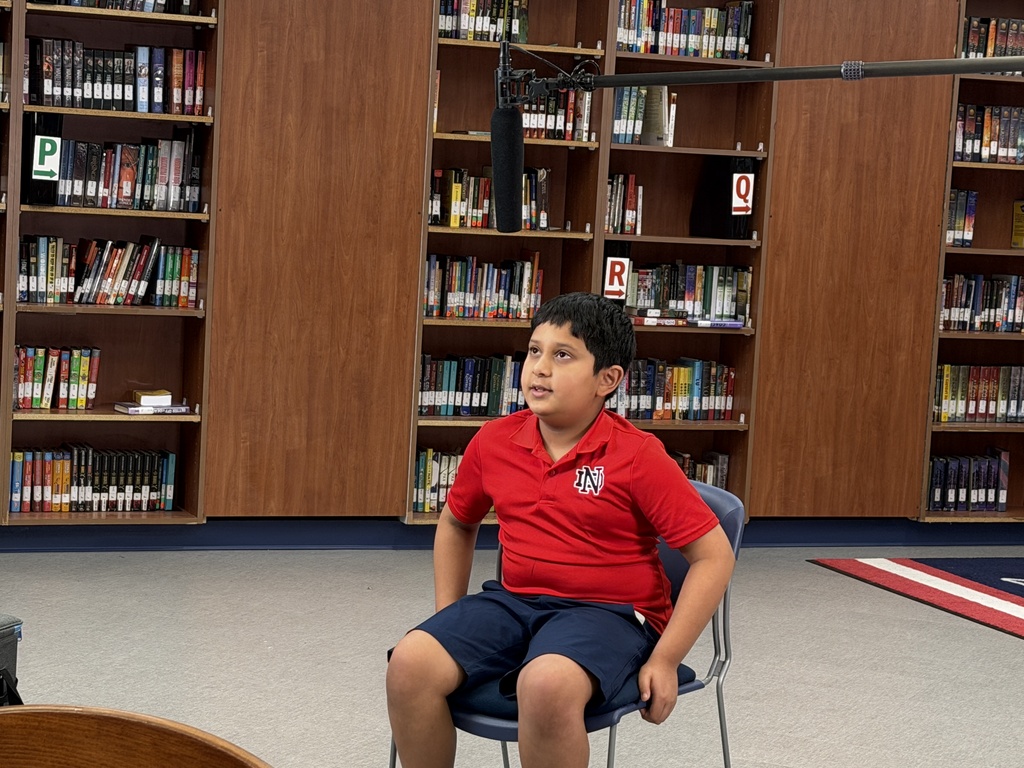 A young boy in a red shirt and blue shorts sits on a chair in a library.