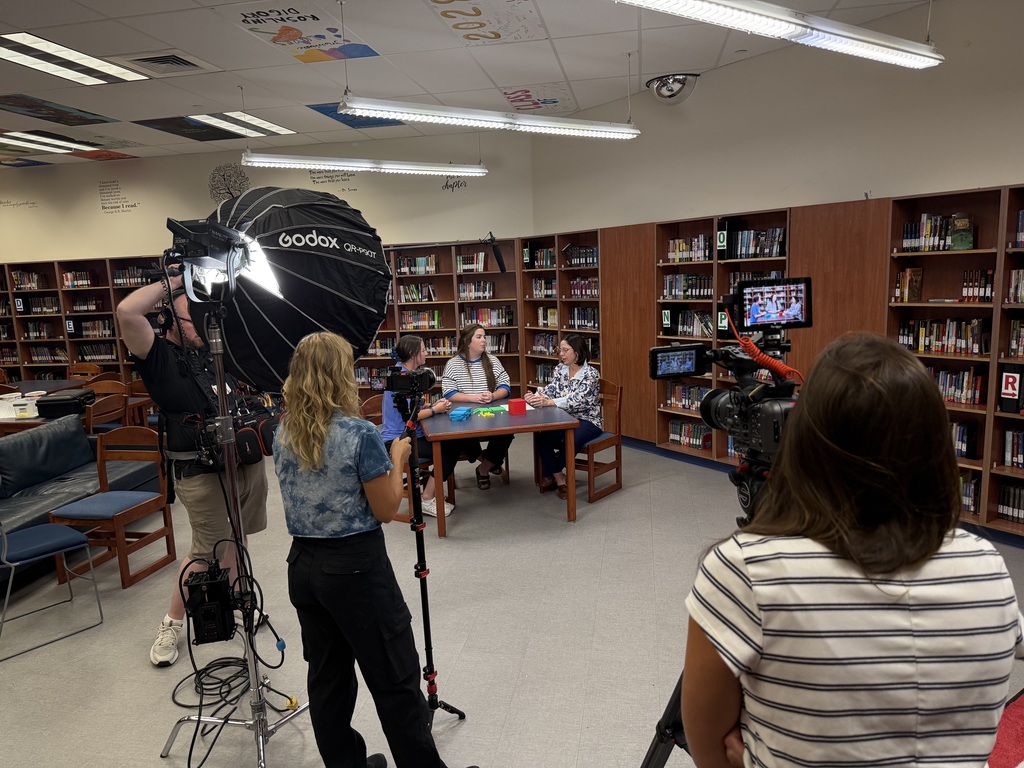 Several people are inside a library, with two women seated at a table and a person holding a camera.