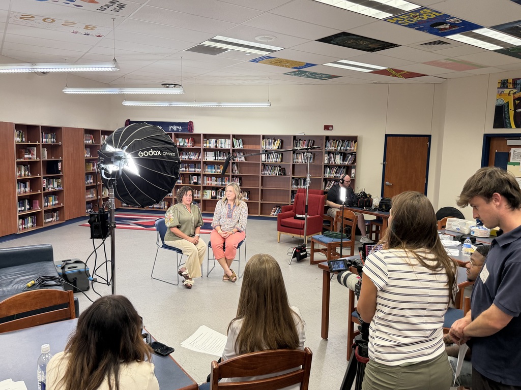 Library room with bookshelves, tables, and chairs. Some individuals sit in chairs, while others stand with cameras.