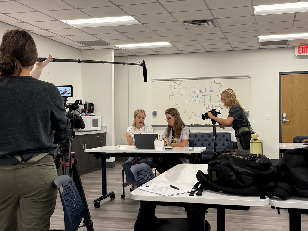 People in a room with desks and chairs, one holding a camera. One person sits at a desk with a laptop.