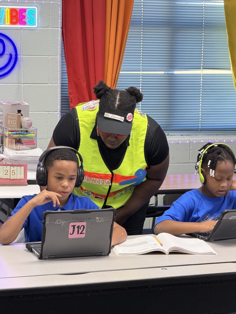 Two children wearing headphones sit at a table with laptops. A woman in a safety vest stands behind them.