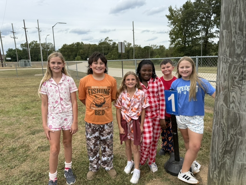 A group of children wearing casual clothing are standing together on grass, with a wooden post on the right.