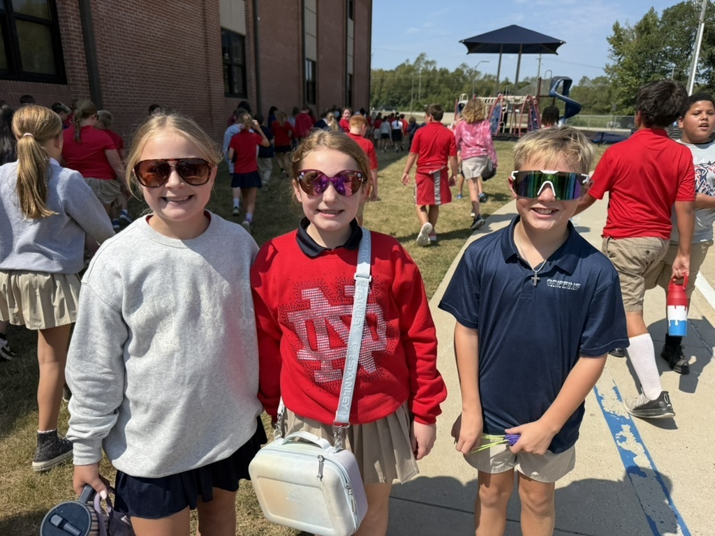 Three children standing, wearing sunglasses, with a school building and several other children in the background.