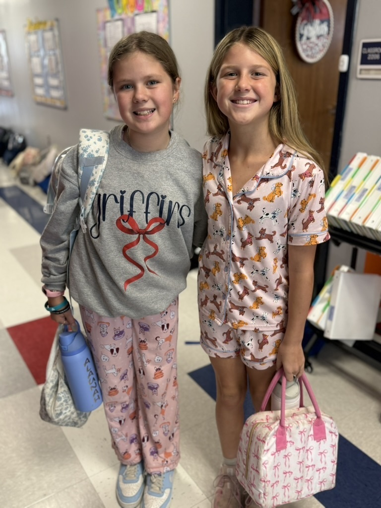Two girls in pajamas, one with a backpack, pose for a photo in a school hallway.