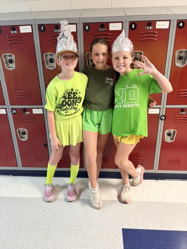 Three children in green shirts and shorts pose in front of lockers. They wear hats, socks, and sneakers.