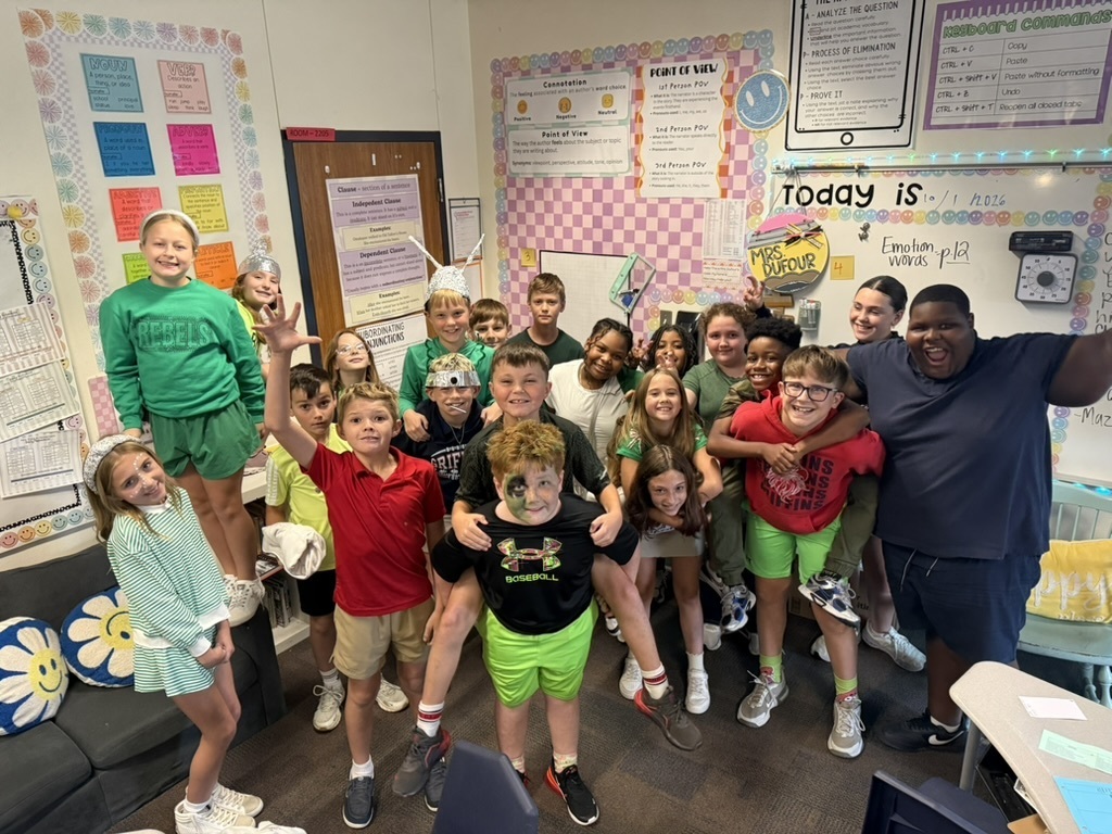 Children and an adult stand in a classroom, raising arms, with colorful walls and furniture.