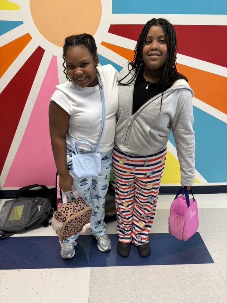 Two individuals stand side by side in front of a vibrant, multicolored wall. Both wear casual clothing.