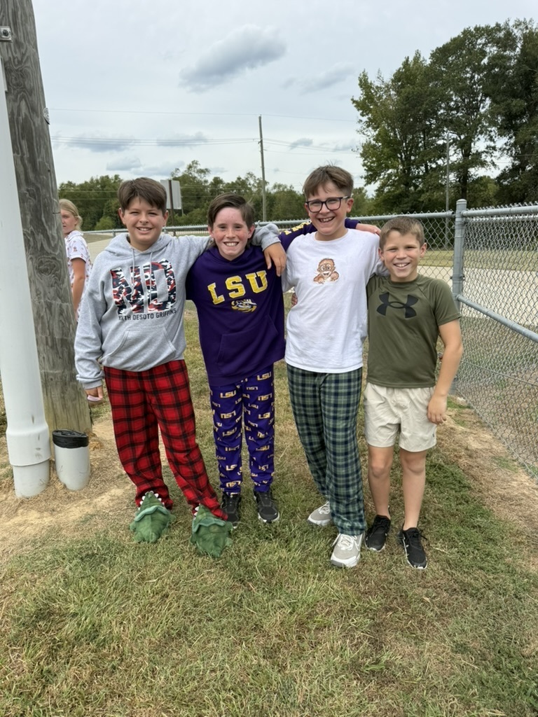 Four kids stand together on grass in front of a fence. They wear different colors of long-sleeve shirts and pants.