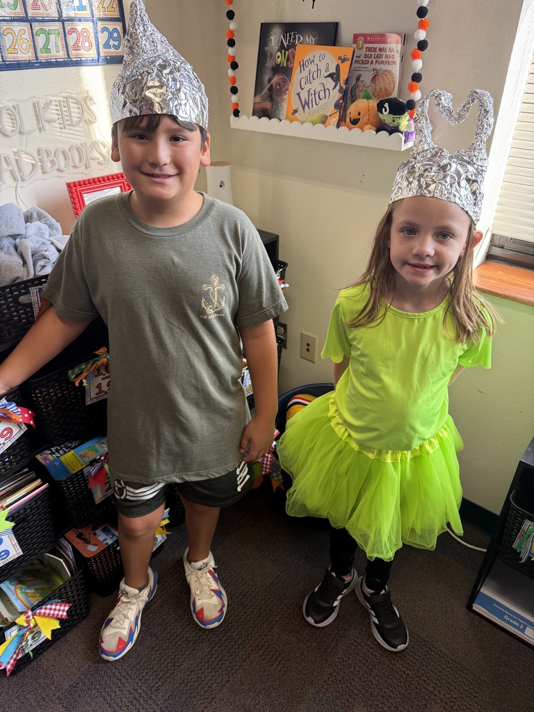 Two children with silver hats, a boy in a green shirt and a girl in a green dress, standing near a shelf filled with books.