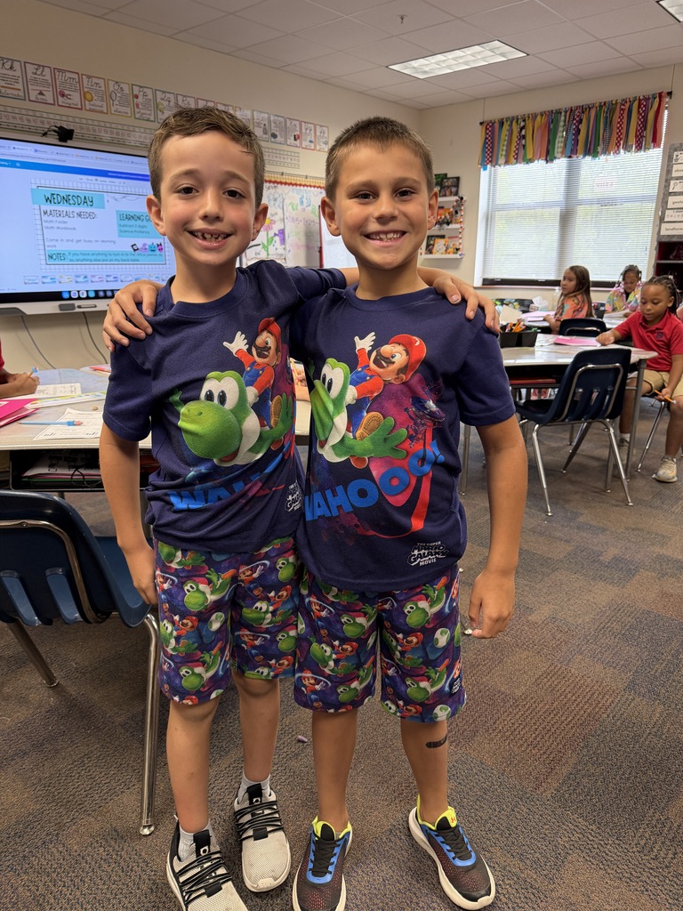 Two boys wearing matching shirts and shorts stand in a classroom, facing the camera. Behind them, tables and chairs are set up.