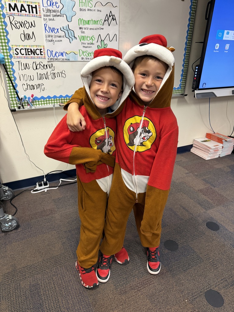 Two children in red and brown costumes stand close together in a classroom with whiteboards and a monitor behind them.