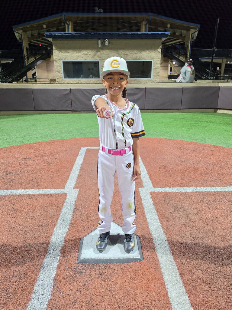 Child standing at home plate on a baseball field, wearing a baseball uniform, hat, and sneakers.