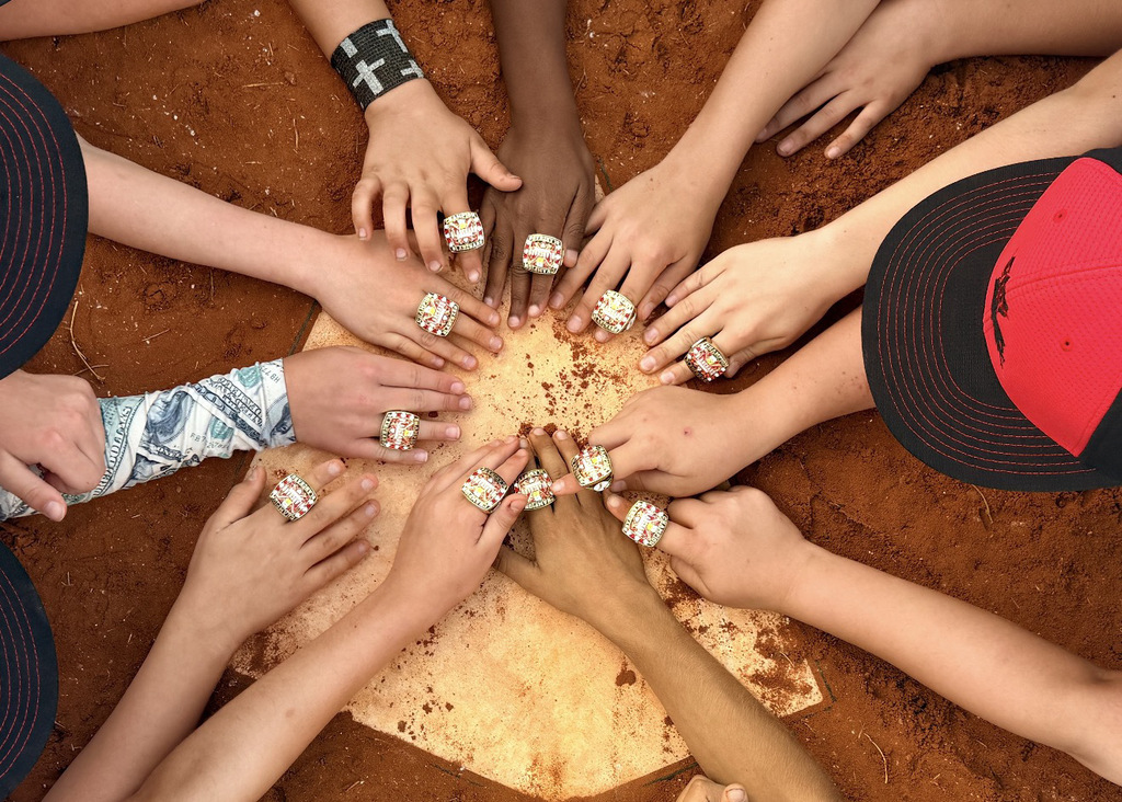 Multiple hands forming a circle around baseballs on a piece of cardboard; baseball caps worn by some individuals.