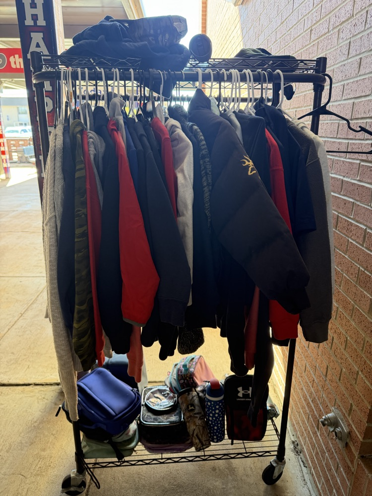 Outdoor rack with hanging clothing, including jackets, hats, and backpacks, next to a brick wall.