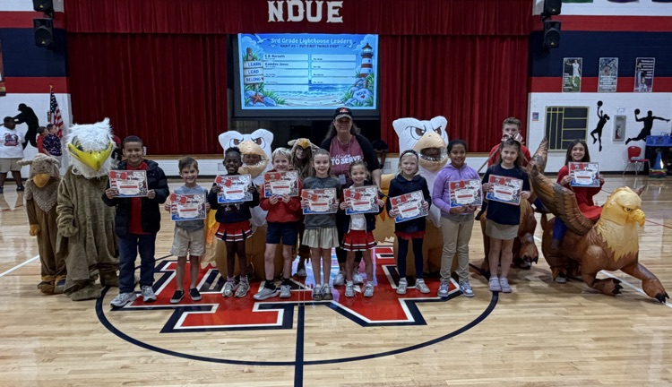 Several children and mascots on a basketball court hold up certificates in front of a red curtain.
