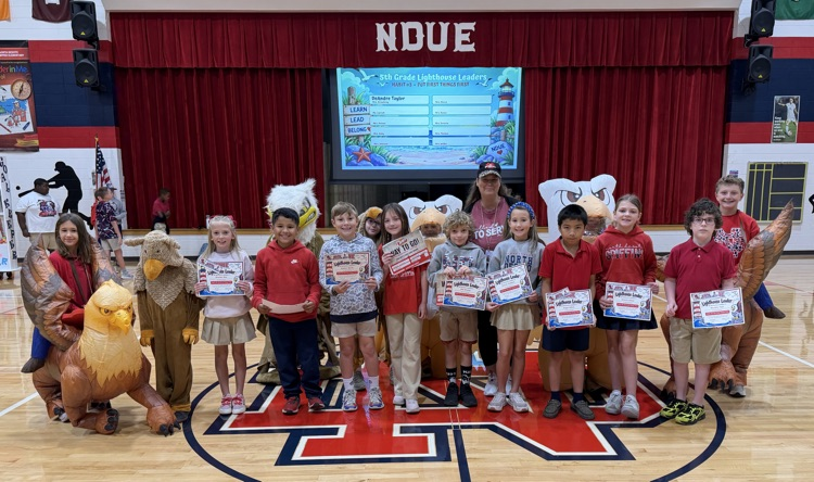 Children in red and white shirts holding certificates in front of a large screen.