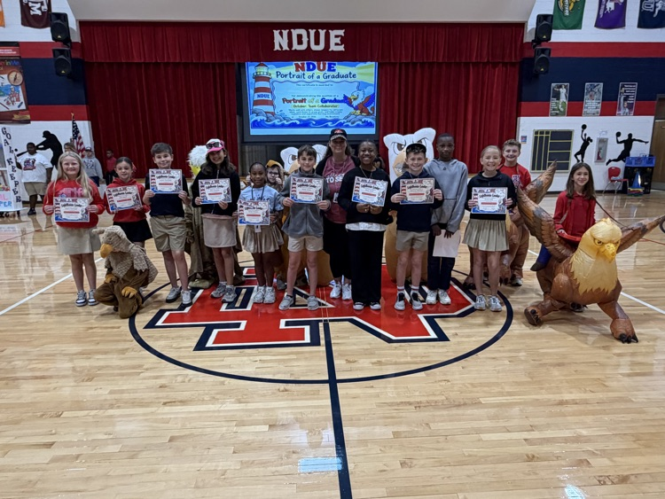 Group of children holding certificates on a basketball court, mascots, large screen at the back.
