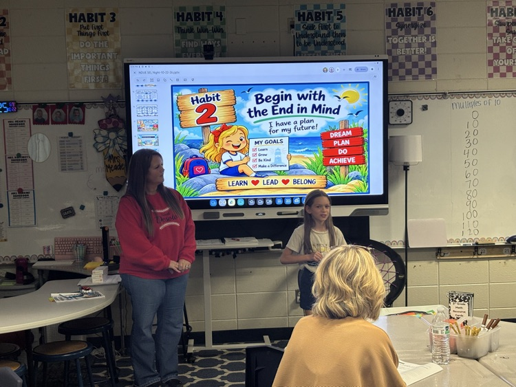 Two females stand before a monitor displaying a screen; one sits at a desk. A woman with blonde hair sits at a table.