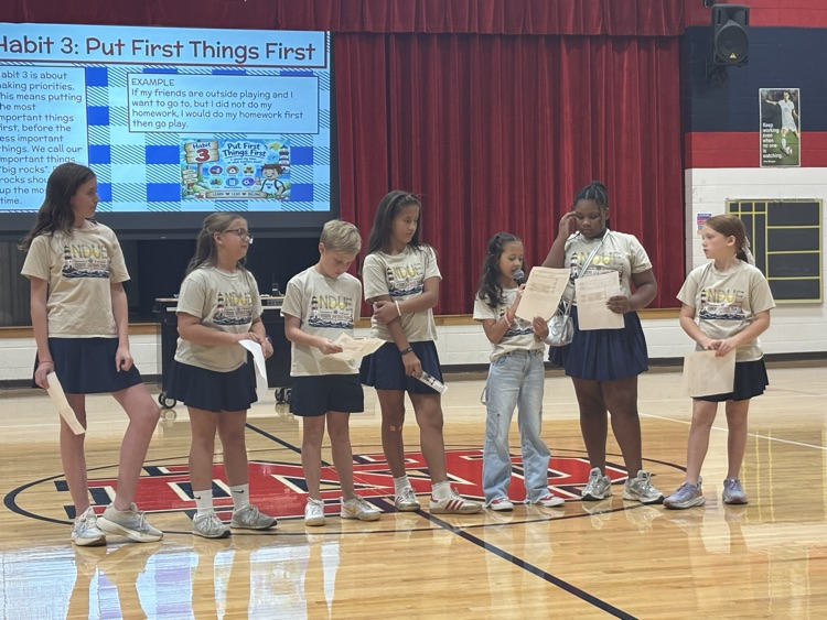 Indoor basketball court with eight children in matching shirts standing in a row holding papers, red curtains in the background, and a projector screen.