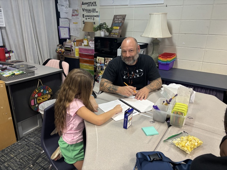 Teacher sitting at a round table writing while a young student sits next to him working on paper.