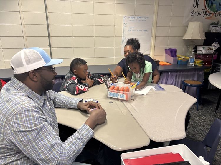 Adult man with hat sits at desk with two children. They write with crayons and hold coloring sheets.